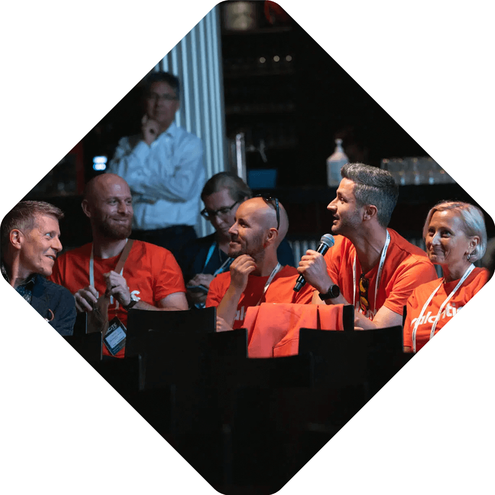 A group of people wearing matching orange shirts sit together at Spryker EXCITE 2025; one person holds a microphone and speaks while others listen.