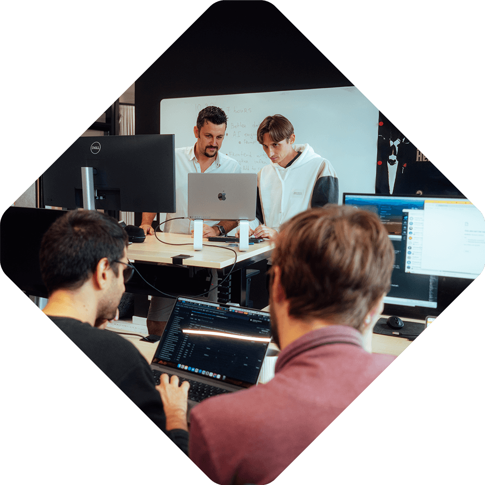 Four men work on laptops in a modern office; two stand by a whiteboard discussing Spryker EXCITE 2025, while two sit at desks facing computer screens.