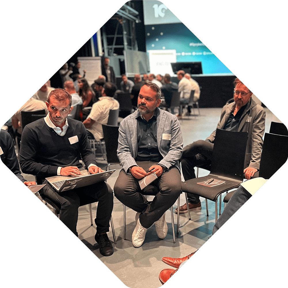A group of men sit in a circle having a discussion at an indoor Spryker EXCITE 2025 event, with others in similar groups in the background.