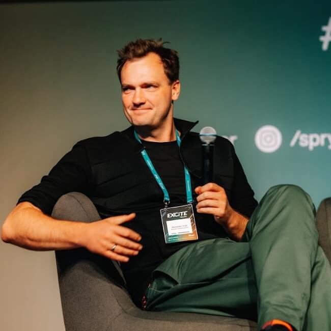 A person with a conference badge sits on a chair, holding a microphone. The background displays social media handles for Spryker EXCITE 2024.