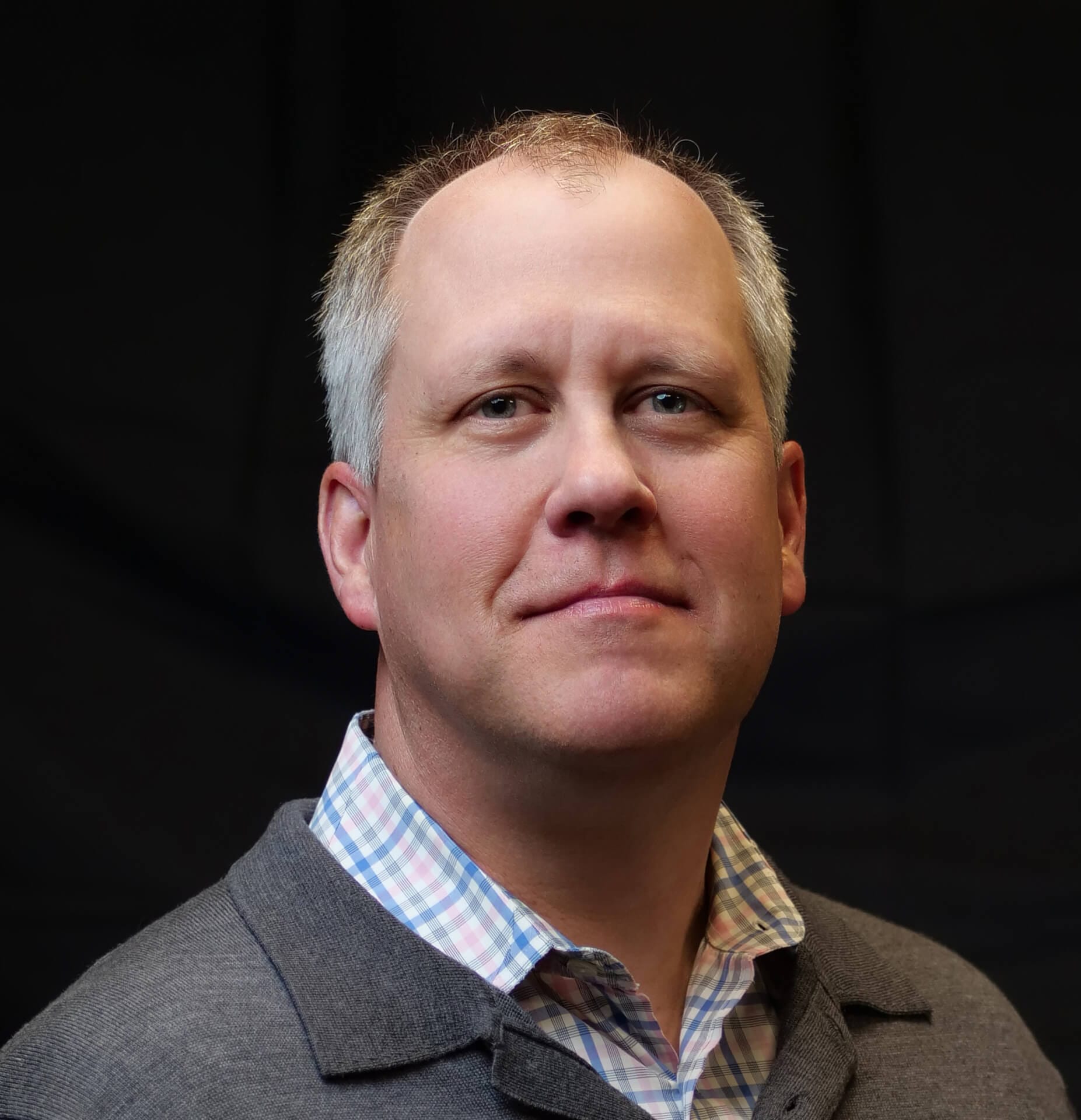 A middle-aged man with short grey hair and a slight smile. He is wearing a grey sweater over a checkered shirt. The background is dark and plain.