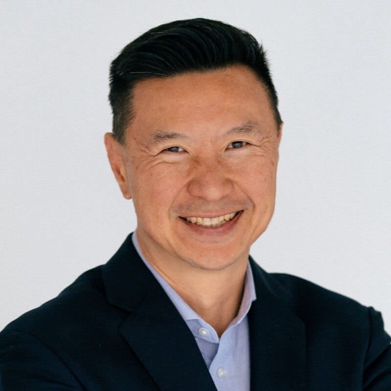 A smiling man with short black hair, wearing a dark jacket and light blue shirt, stands against a plain white background, ready to attend Spryker EXCITE 2024.