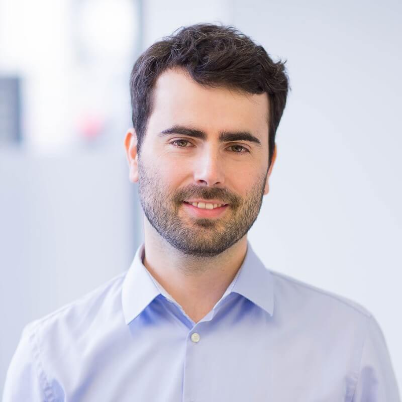 A man with dark hair and a beard wearing a light blue button-up shirt stands against a plain background, smiling slightly, exuding the enthusiasm of Spryker EXCITE 2024.