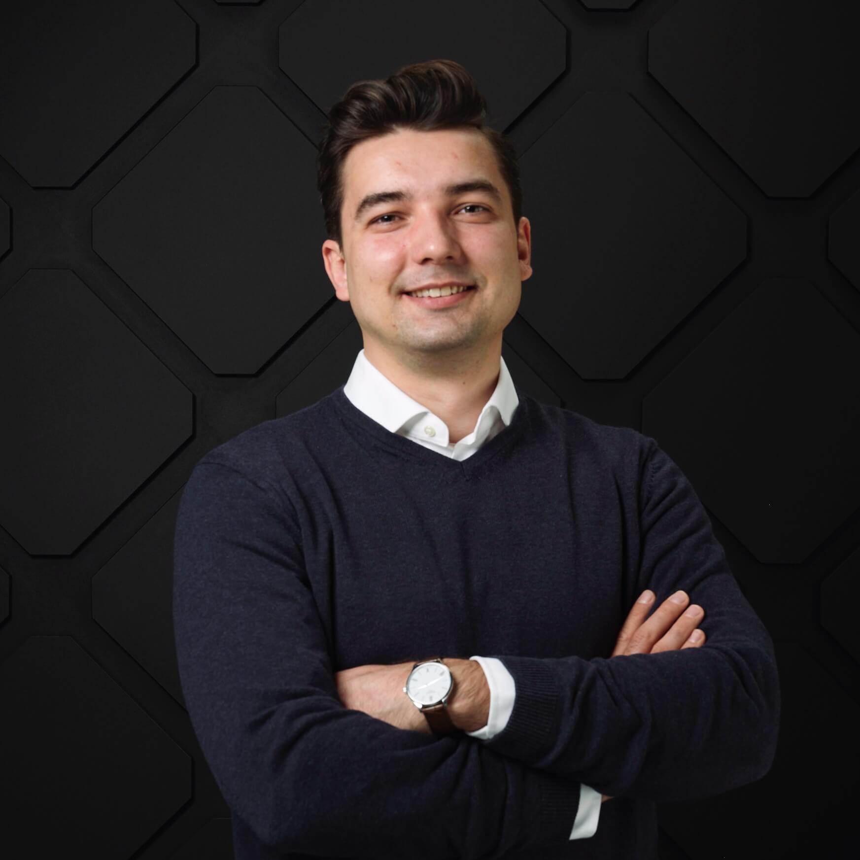 A man with short dark hair, wearing a dark sweater over a white shirt and wristwatch, stands against a dark, geometric-patterned background at Spryker EXCITE 2024, smiling with arms crossed.