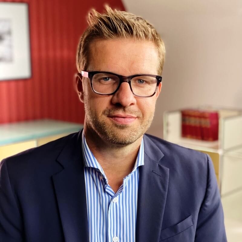 Man with glasses and short blond hair wearing a blue blazer and striped shirt, standing in a room with a red wall and bookshelf, preparing for Spryker EXCITE 2024.