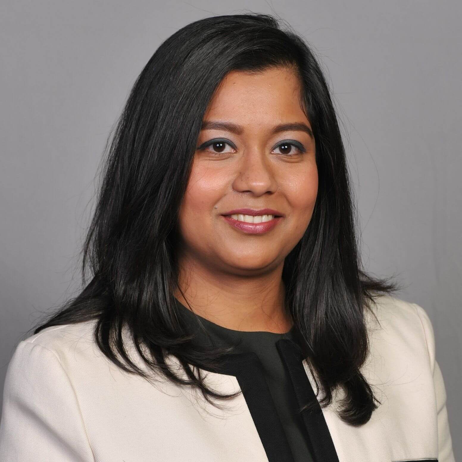 A woman with long dark hair smiles confidently at the camera. She is wearing a cream-colored blazer over a dark top. The background is a plain gray.