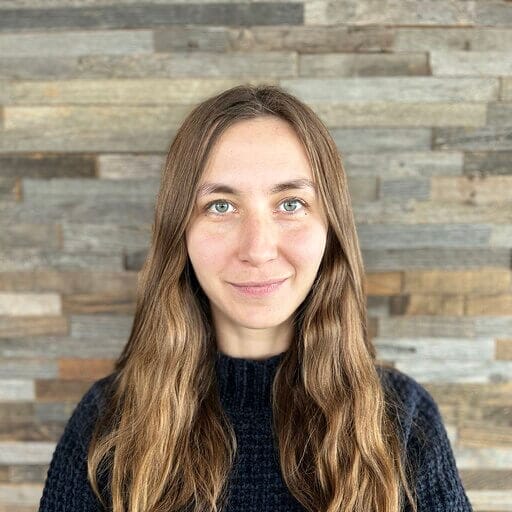 A woman with long, wavy hair and a slight smile stands in front of a wooden wall at Spryker EXCITE 2024. She is wearing a dark top, and the background consists of various shades of wooden planks.