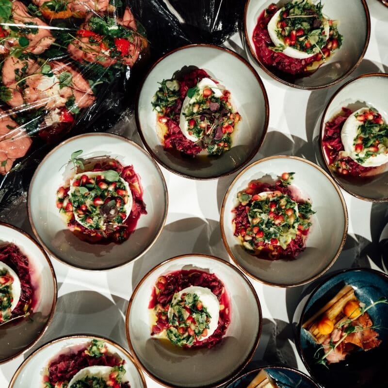 Several bowls filled with a colorful salad featuring greens, pomegranate seeds, and white cheese are arranged on a table next to assorted covered dishes, creating a vibrant setting perfect for Spryker EXCITE 2025.