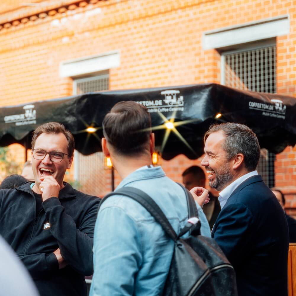 Four men stand outside near a coffee cart at Spryker EXCITE 2025, engaged in conversation and laughter, with a brick building and a black awning in the background.