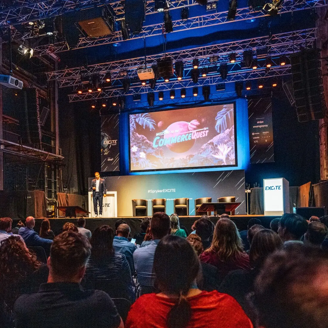 A speaker is presenting on a stage with a large screen displaying "CommerceQuest" to a seated audience in a dimly lit venue. The background includes stage lights and structural elements, and the banners on either side of the stage read "EXCITE.