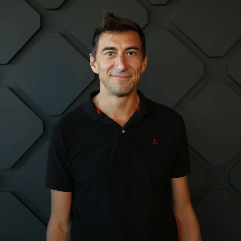 A person with short dark hair wearing a dark polo shirt is standing in front of a textured, geometric-patterned dark wall. The person is smiling and looking directly at the camera.