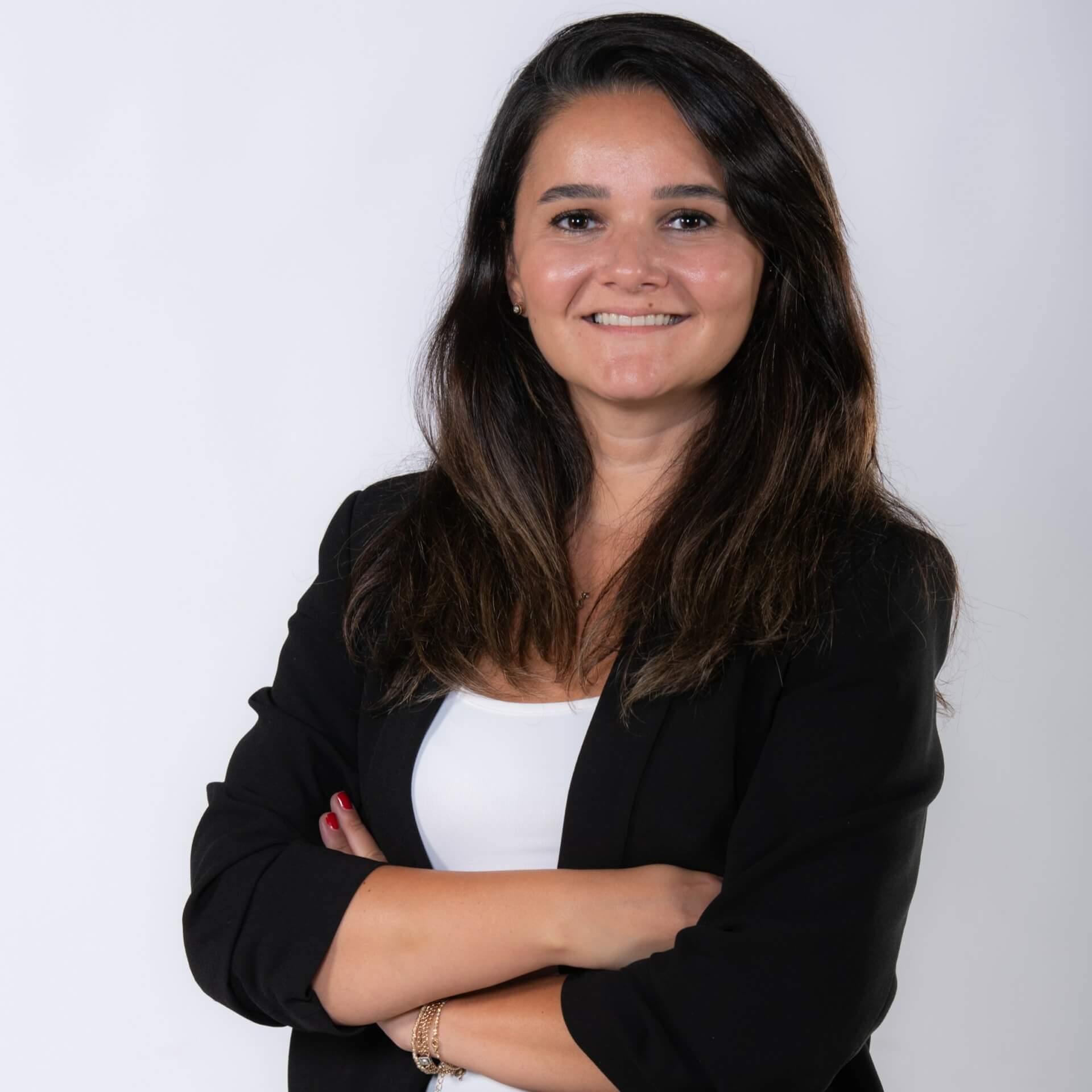 A person with long dark hair, wearing a black blazer and white top, is smiling at the camera with arms crossed in front of a plain white background, ready to represent Spryker EXCITE 2024.