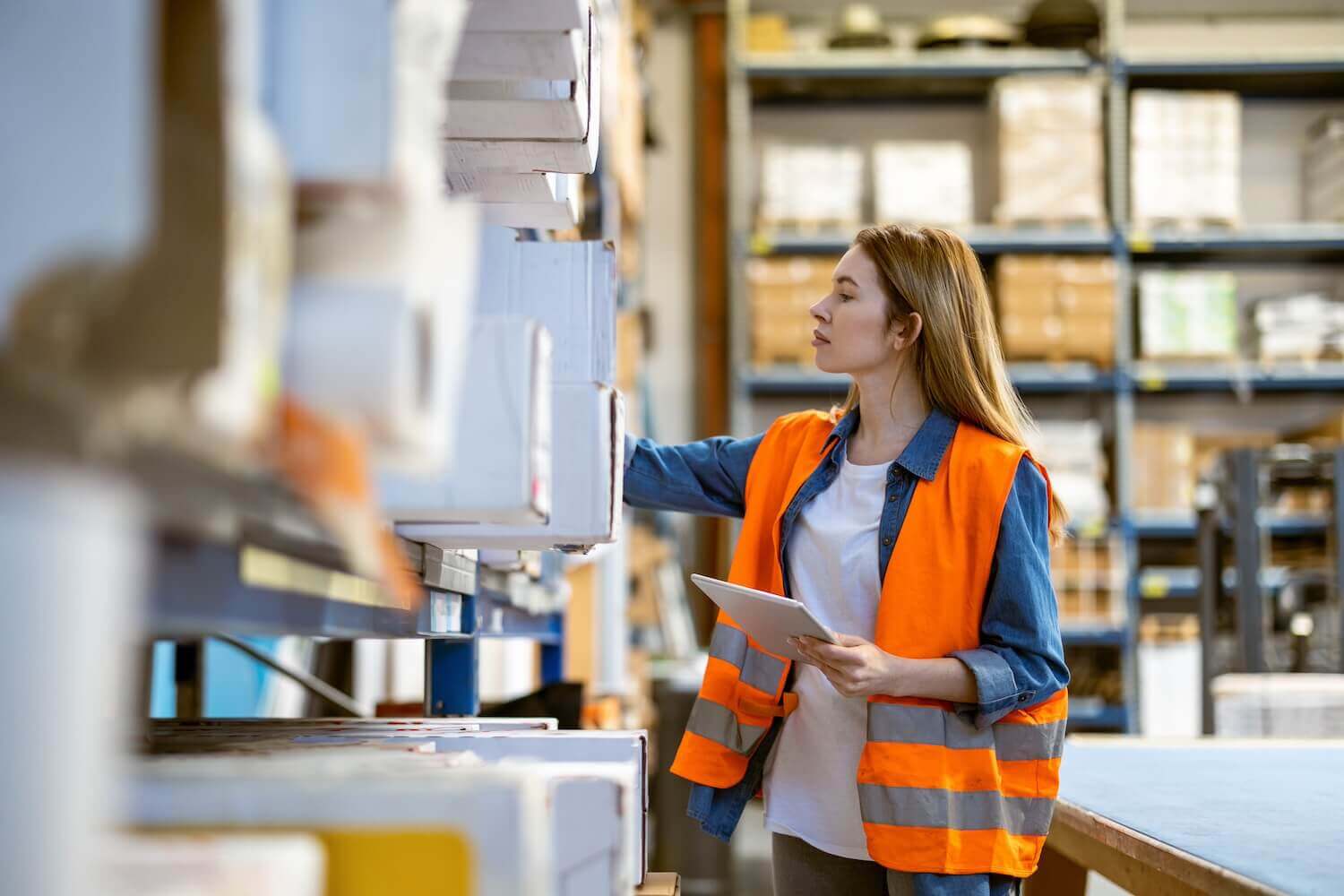 A woman wearing an orange safety vest and holding a tablet is standing in a warehouse, reaching for an item on a shelf. The background shows shelves stocked with various boxes and items.