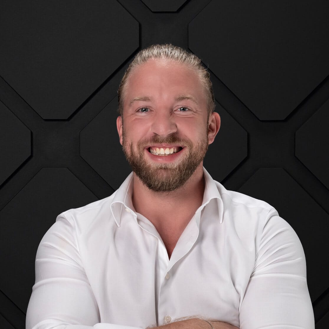 A smiling person with a beard and slicked-back hair, wearing a white dress shirt, is standing against a backdrop with a geometric black pattern. Arms are crossed in front.