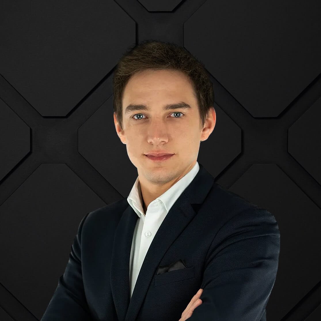 A man in a dark suit and white shirt stands confidently with his arms crossed. He has short brown hair and is set against a dark, geometric background.