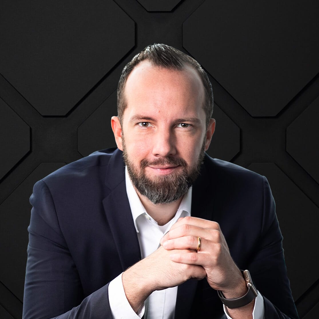 A man with short hair and a beard, dressed in a dark suit and white shirt, sits with his hands clasped in front of him against a dark, hexagonal-patterned background. He has a focused expression.