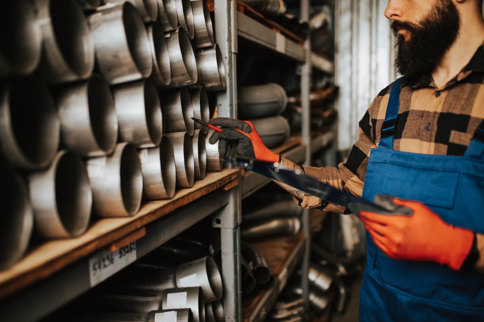 A person wearing an orange plaid shirt, blue overalls, and orange gloves is picking a metal pipe from a shelf in a workshop. The shelves are filled with various metal pipes and fittings.