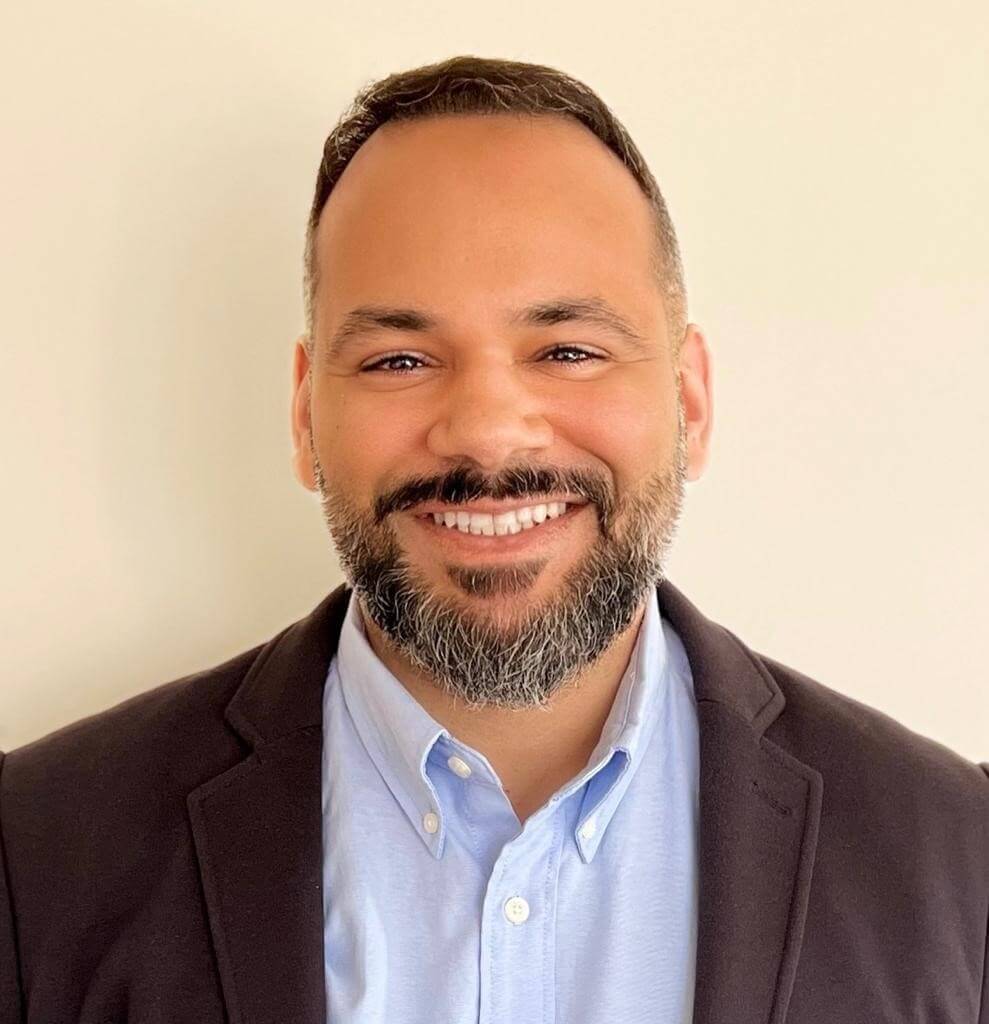 A man with short dark hair and a beard is smiling. He is wearing a blue dress shirt and a dark blazer. The background is plain and light-colored.