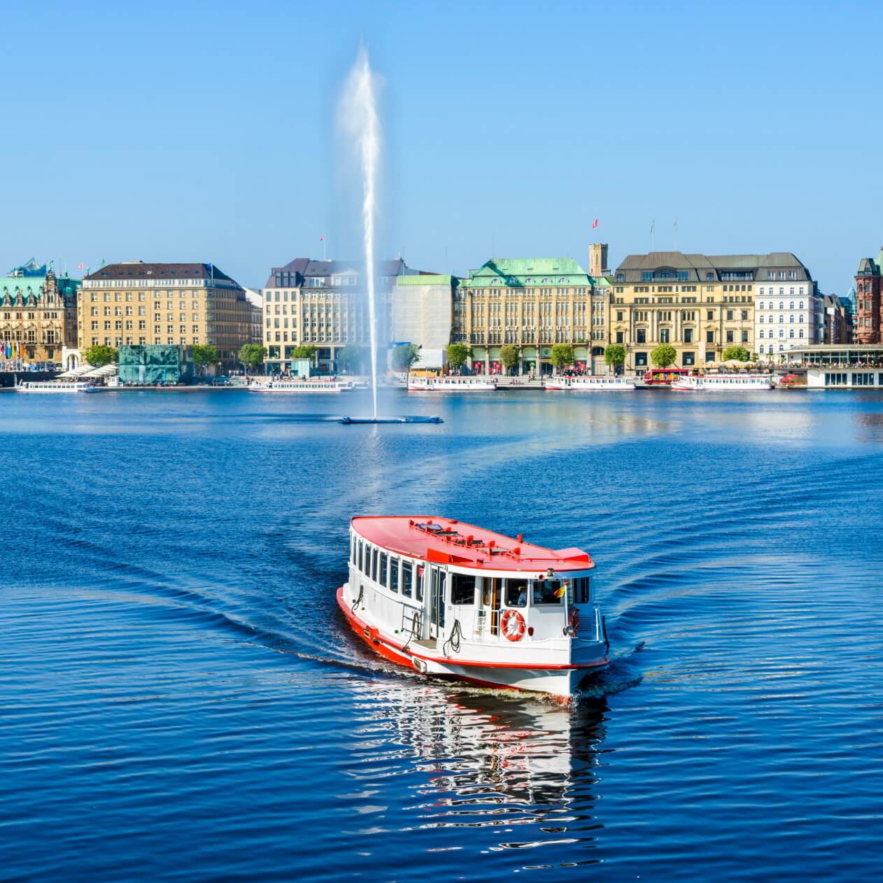 A white and red boat sails on a calm lake with ripples trailing behind it. In the background, a cityscape with historic buildings lines the waterfront, and a tall water fountain sprays into the air against a clear blue sky.