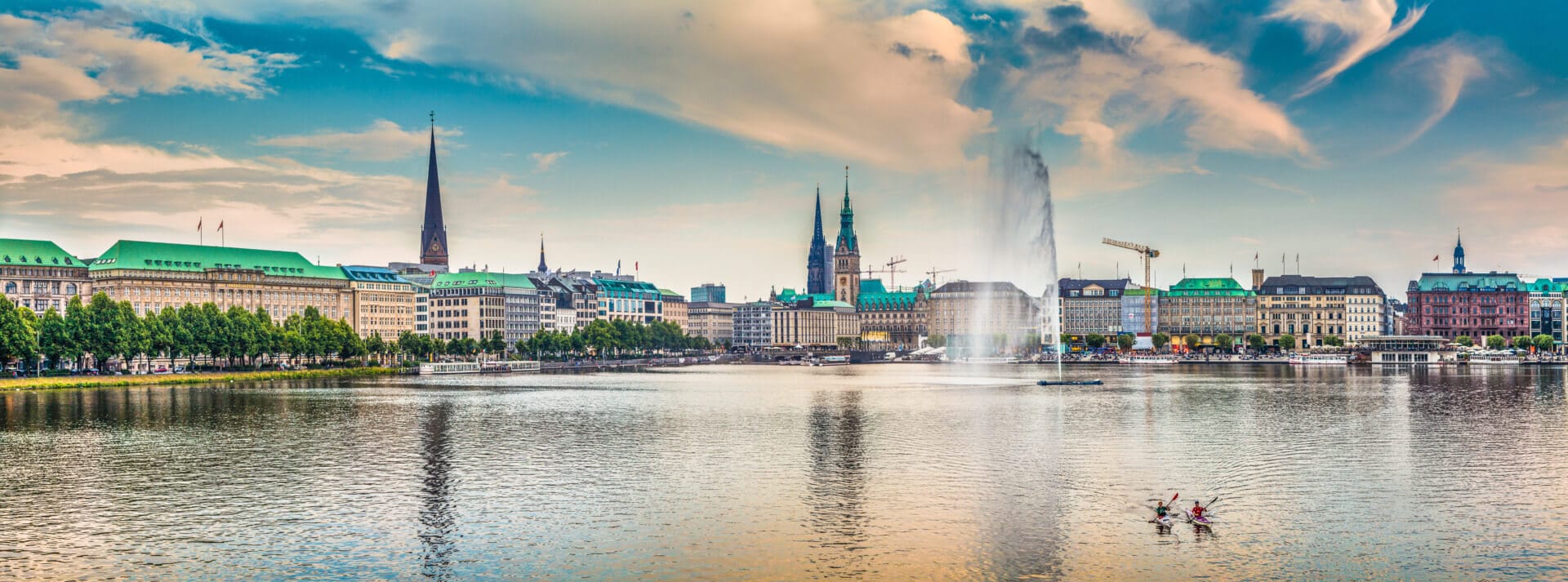 Panoramic view of the Binnenalster, the inner portion of the Alster lake in Hamburg, Germany. The skyline features historic buildings, church spires, and a large fountain. Two small boats with rowers are seen in the foreground on the water. The sky is partly cloudy.