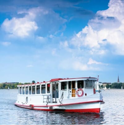 A white and red riverboat is anchored on a calm waterway against a backdrop of a partly cloudy blue sky. Sailboats can be seen in the distance, and lush greenery lines the shore under the bright daylight.