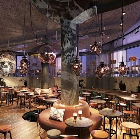 A stylish bar interior featuring modern decor with pendant lights hanging from the ceiling, bar stools, and round tables. The space has a focal column with artistic designs and warm, ambient lighting. Two filled glasses sit on a central round table.