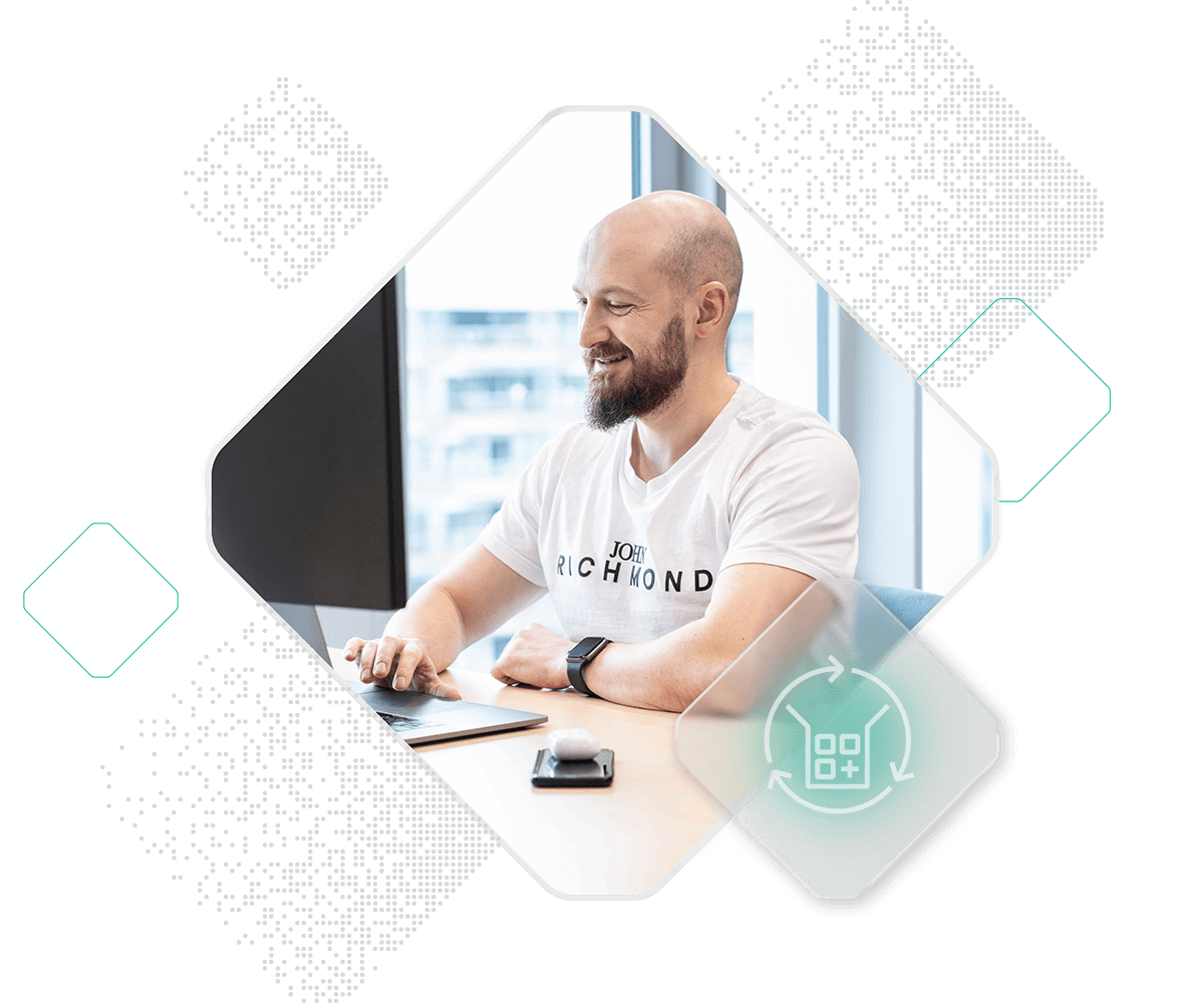 A man with a beard is sitting at a desk, using a laptop. He is wearing a white t-shirt with text on it and an activity tracker on his wrist. The background contains pixelated digital patterns and abstract shapes.