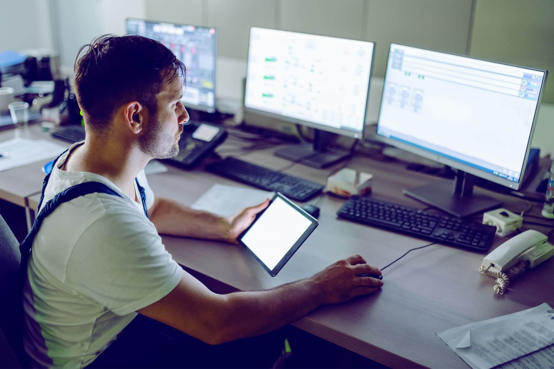 A man sits at a desk with multiple computer monitors and holds a tablet. He appears to be focused on the screens, which display various data and graphs. The environment suggests a high-tech or control room setting. Documents and office supplies are scattered on the desk.