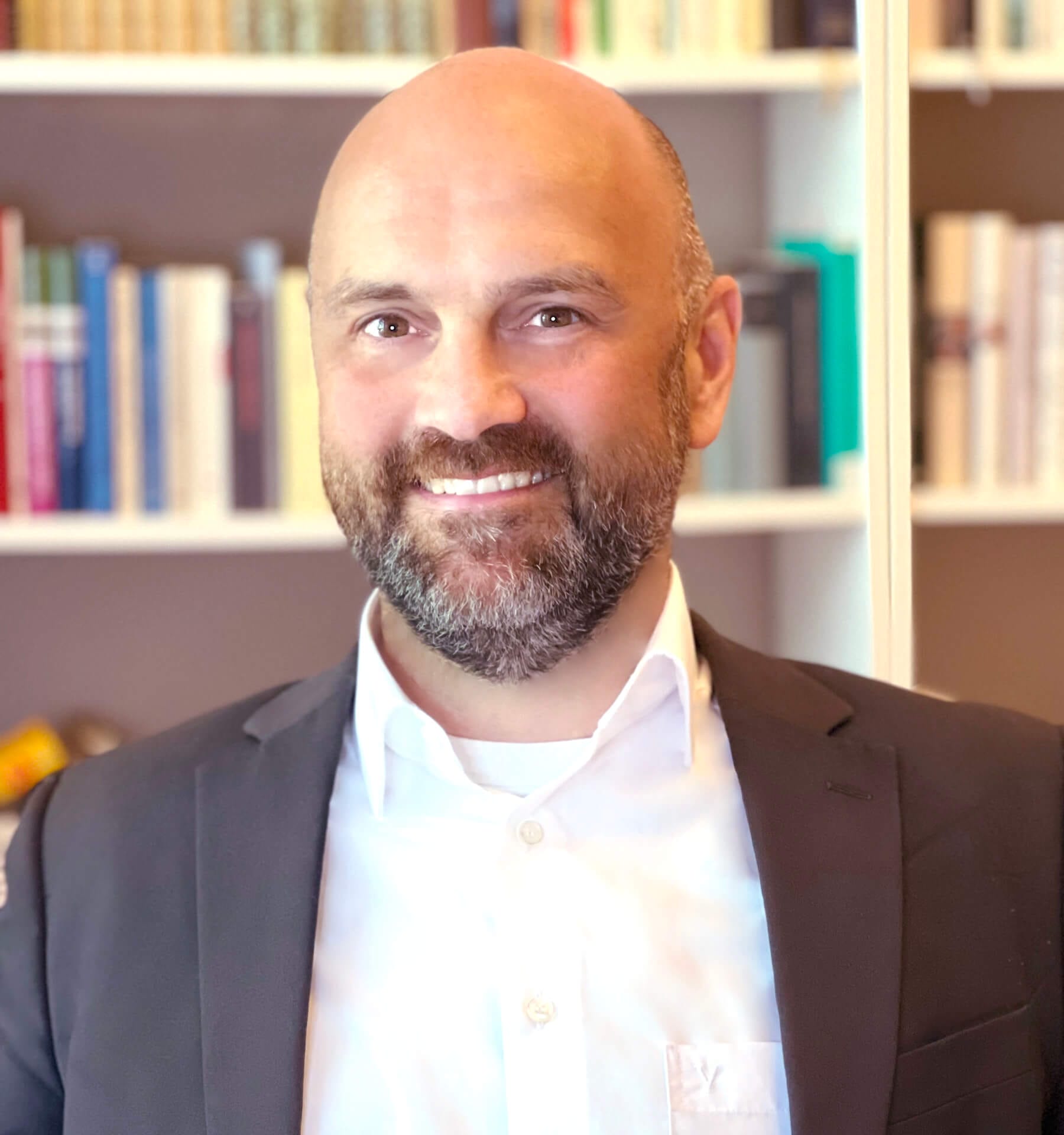 A smiling man with a bald head and a beard is wearing a white dress shirt and dark blazer. He is standing in front of a bookshelf filled with various books.