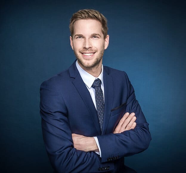 A man with light brown hair and a beard is wearing a navy blue suit and tie, posing against a dark blue background. He is smiling and has his arms crossed in front of his chest.