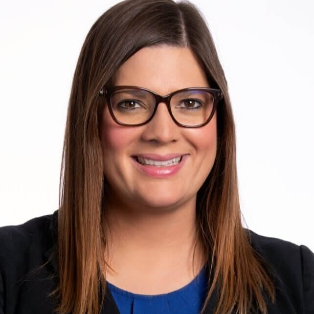 A woman with long brown hair, wearing glasses with dark frames, smiles while wearing a blue top and a black blazer. She is posed against a plain white background.