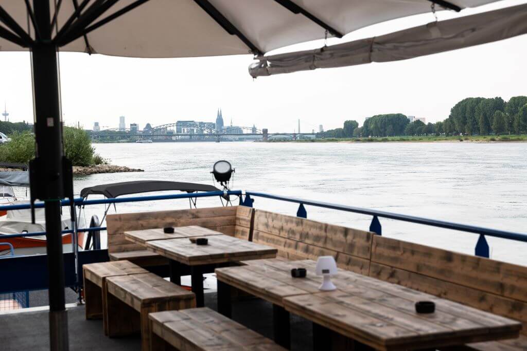 A riverside seating area under an umbrella with wooden tables and benches. A cityscape with a bridge and buildings can be seen in the background across the calm river, perfect for watching the upcoming OMR 2024 amidst serene surroundings.