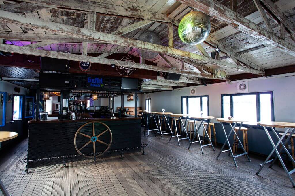 A rustic bar area with exposed wooden beams, a central counter, high tables and stools, a disco ball on the ceiling, and various decorations on the walls captures the essence of OMR 2024's vibrant social vibe.