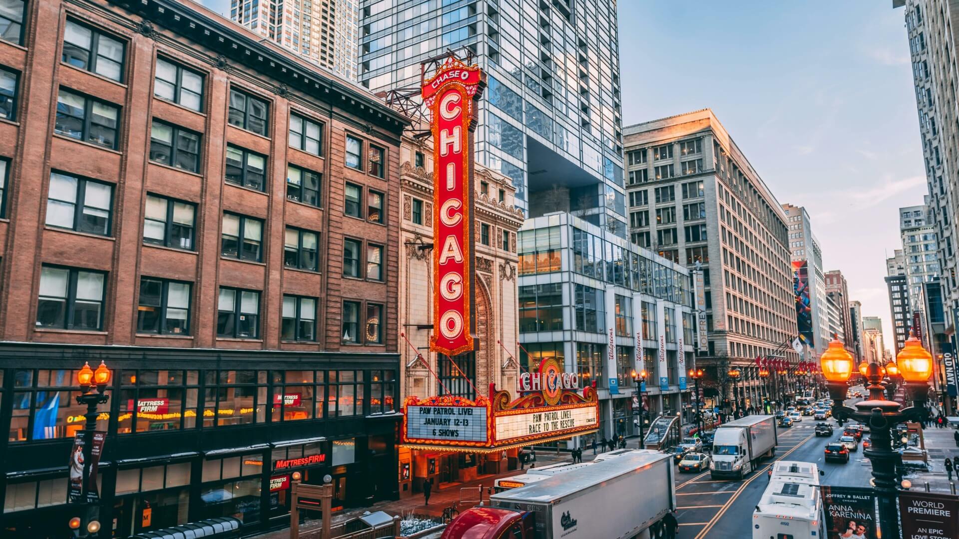 City street view featuring the Chicago Theatre with its iconic neon sign and bustling traffic, capturing the vibrant energy of downtown Chicago. Mid-rise buildings line the street under a clear blue sky, as banners for CES 2024 hint at exciting events on the horizon.