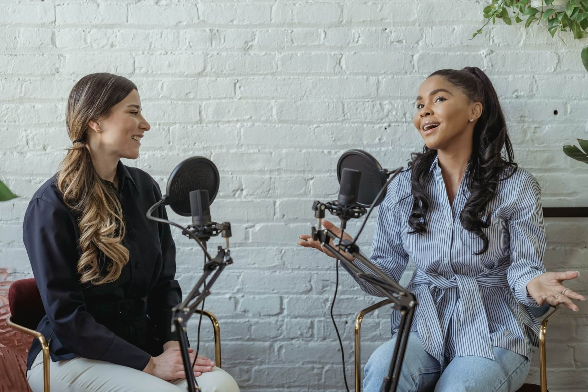 Two women sit in front of microphones, engaged in a conversation about the upcoming OMR 2024 event. They face each other with a white brick wall in the background.