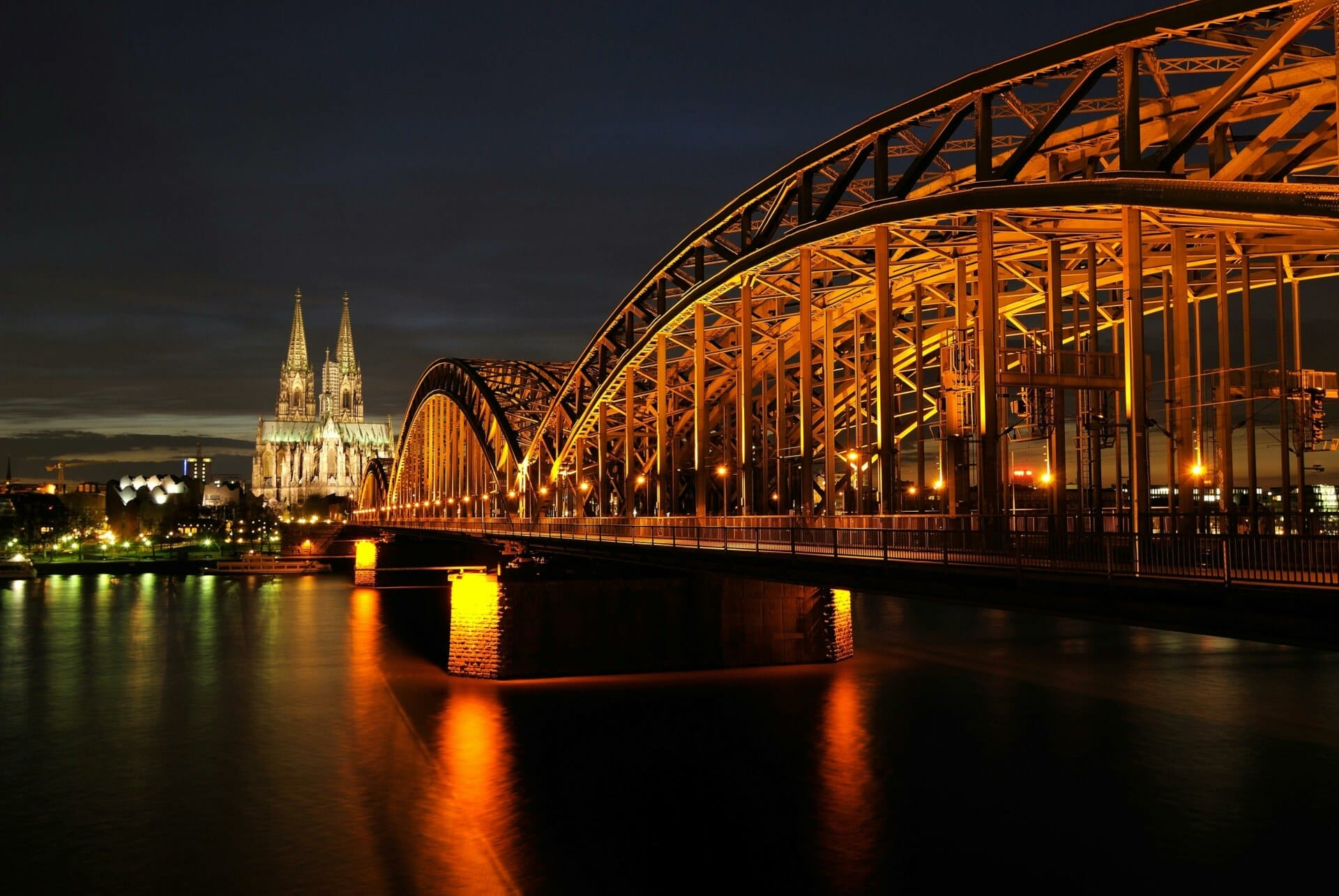 A brightly lit, ornate bridge stretches across a reflective river at night, with a large, illuminated cathedral visible in the background. The scene elicits dreams of Omr 2024, where history meets modernity in a stunning display.