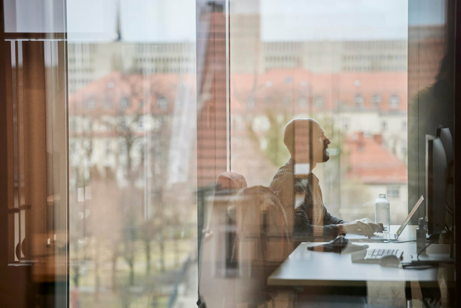 A person is seated at a desk in an office, using a laptop. Reflections of the window and the outside view, which includes buildings and trees, are visible on the glass. Subtly placed on a shelf nearby are certificates showcasing AWS Competencies, adding a touch of professionalism to the scene.