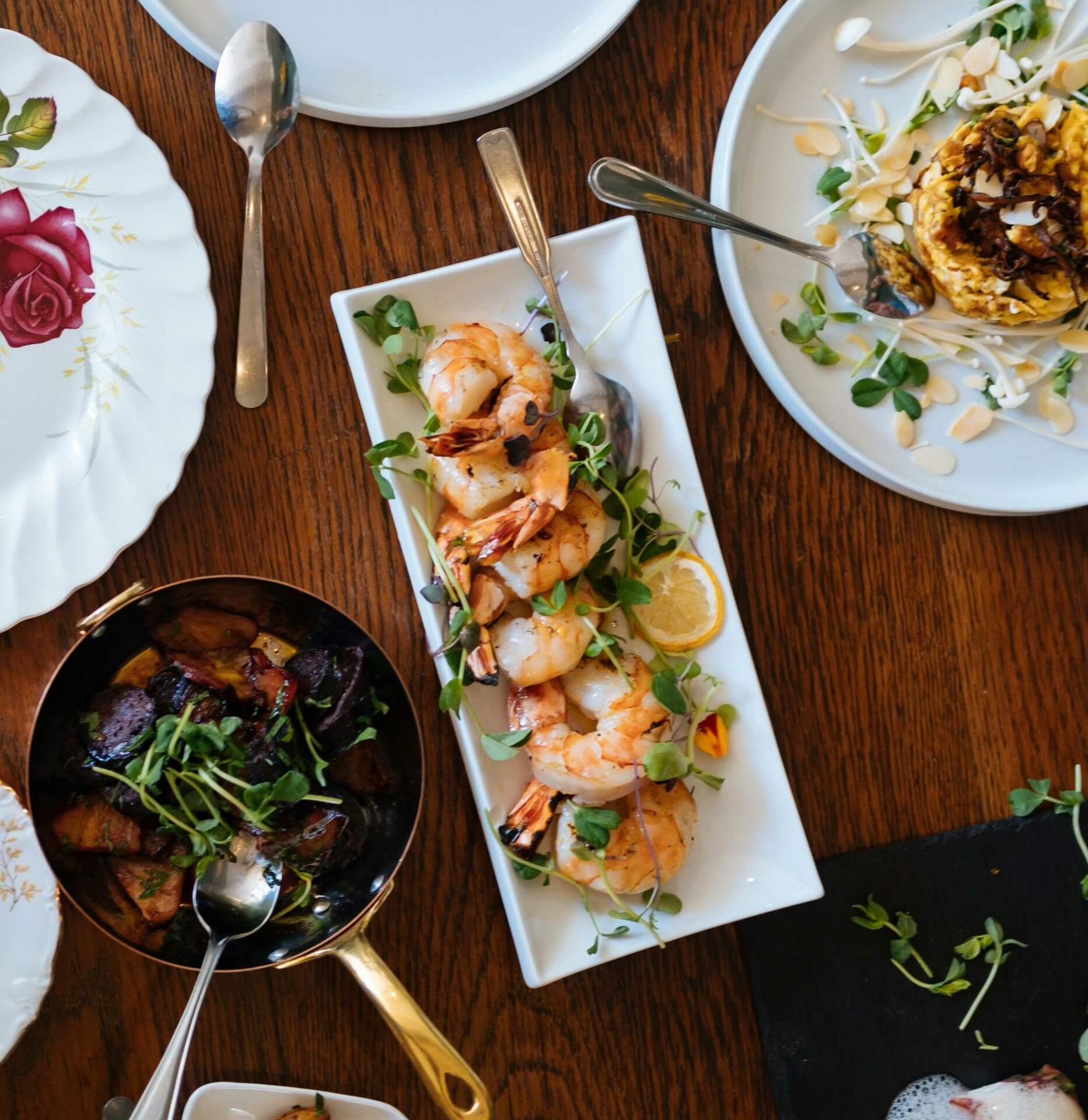 An overhead view of a table with various dishes, including grilled shrimp on a rectangular plate, roasted potatoes, and a corn dish garnished with herbs. It's like preparing for B2B Online Europe 2024—a feast for the eyes and taste buds alike.