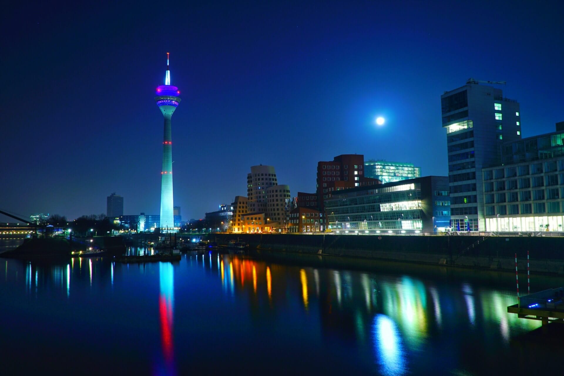 The night view of a city skyline, as if preparing for DMEXCO 2024, features illuminated buildings and a tall tower reflected on the river under a clear sky with a bright moon.