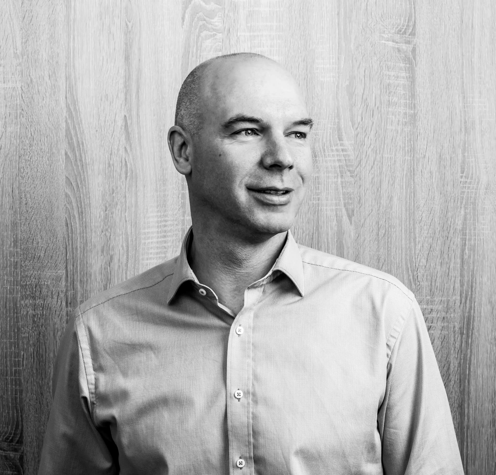 A bald man in a light-colored button-up shirt stands in front of a wooden wall, looking to the right, seemingly lost in thought about an upcoming side event at B2B Online Europe 2024. Black and white photo.