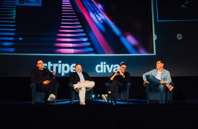 Four people are seated on stage, engaged in a panel discussion about Spryker. A colorful projection is visible behind them, enhancing the lively atmosphere.