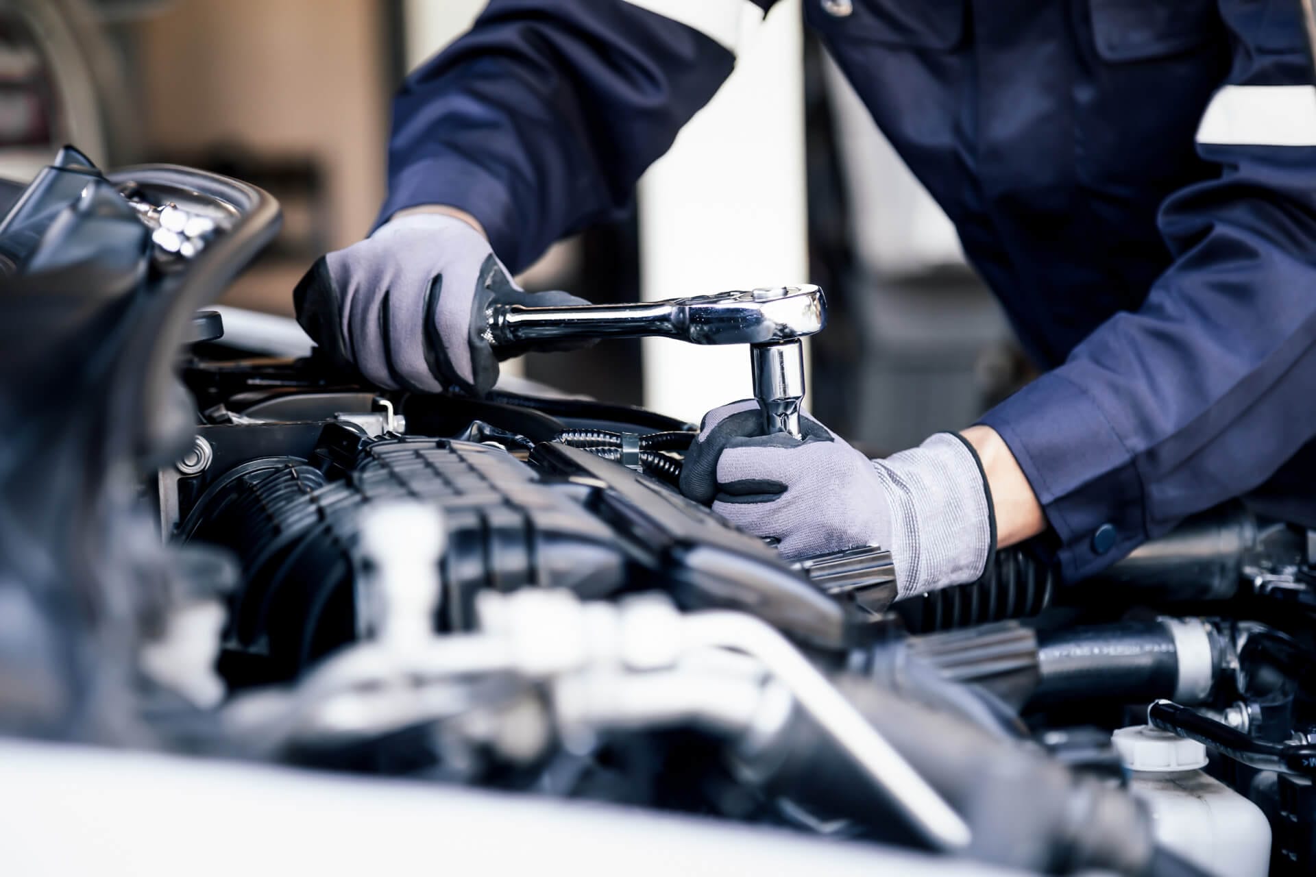 A mechanic in a blue uniform expertly uses a ratchet wrench to work on a car engine, wearing gloves to ensure both safety and precision—a testament to the quality of consumer goods in automotive maintenance.